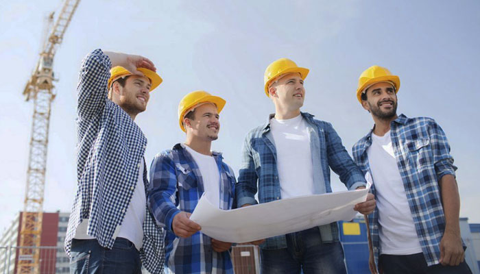 Patru muncitori pe șantier, poartă căști galbene de protecție și cămăși în carouri albastre, privesc optimiști și țin planuri de construcție, pe fundal cu macarale